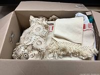 Box containing vintage textiles including lace doilies and fringed linen pieces.