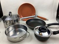 Full set of pots and pans arranged including stainless steel, black non-stick, and copper-colored items.