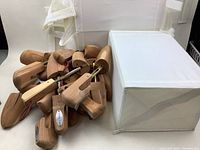 Wide shot of a pile of wooden shoe stretchers with attached metal rods and maroon rope handles, alongside four white vinyl shoe holders.