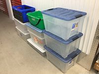 Photo showing nine assorted plastic storage bins lined up against a white wall. Various colors include clear with blue lids, solid blue with black base, green with black handle, and gray bins without lids.