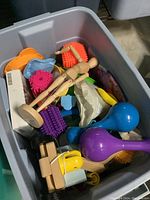 Photo showing a plastic bin filled with a variety of kids toys including wooden mannequins, sensory brushes, plastic maracas, yellow saw, and plastic toys.