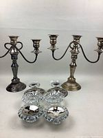 Two tarnished metal three-arm candelabra candle holders with curved arms and four small clear glass candle holders with scalloped edges arranged on a white surface.