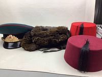Four vintage hats arranged against a plain background showing a Soviet military officer visor cap, a fur Ushanka, and two red fezzes.