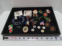 Photo of assorted Christmas-themed jewelry items arranged on a black surface, showing pins, a bracelet, earrings, and pendants, with a measuring tape for scale.