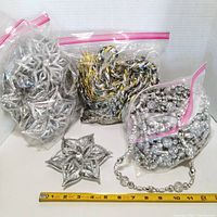 Clear photo showing zip bags with silver star ornaments, silver bead garland, gold and silver twisted garland, and loose silver bead decorations, with a measuring tape for size reference.