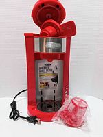 Red BELLA Dual Brew Single Serve Coffee Maker with open lid showing brewing chamber and attached power cord, accompanied by an adapter in plastic packaging.