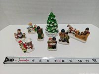 Nine ceramic Christmas village figurines including groups of people, a Christmas tree, and horses with sleigh, arranged in front of a white background with a measuring tape showing size.
