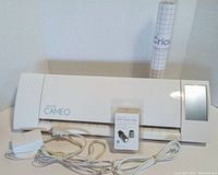 Silhouette Cameo 3 cutting machine front view with touchscreen powered on, power cords, extra blade, and cutting mat rolled up with Cricut branding.