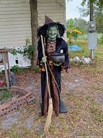 Front view of 5-foot tall Gemmy witch outdoor decoration with broom and cauldron, dressed in black and purple, standing on grass near a building