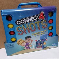 Front of blue plastic Connect 4 Shots board game in open packaging showing game layout, colorful balls, and game handle.