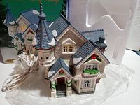 Front and left side view showing detailed Victorian-style roof with turret and porch, electric cord for illumination, sitting in original styrofoam insert with box in background