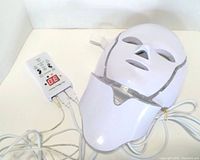 White plastic facial LED skin therapy mask with eye, nose, and mouth openings, adjustable strap, and connected cords with remote control showing settings.