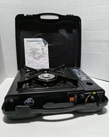 Portable single burner camping stove open in hard black plastic carrying case with instruction sheet included.