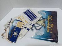 White and blue prayer shawl with gold embroidery and matching decorative bag with winged design, with tags and packaging.