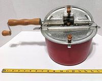 The Genuine Whirley Pop aluminum stovetop popcorn maker with wood hand crank shown on a flat surface next to a ruler for size reference.