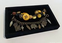 Two vintage charm bracelets laid in a black display tray, showing the full length and details of the charms including an elephant and leaf charms on one and mostly button charms on the other.