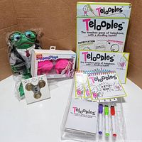 Photo showing set of items: Wallstreet Hubble frog puppet with business outfit, two Teloodles game boxes including sketchbook and markers, pink furry headphones, and metallic fidget spinner.