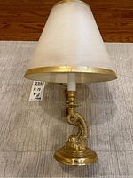 Full view of the table lamp showing the brass fish base and beige fabric shade with gold accent trim.