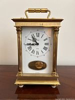 Front view showing the clock face with Roman numerals and decorative hands, brass columns, and handle on top.