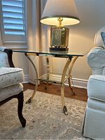 Side view of the brass side table placed between chairs, showing the beveled glass top and lower shelf with a lamp on top.