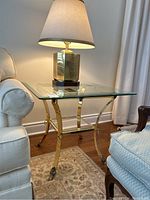 Side view of brass side table positioned between upholstered chairs with a lamp on top, showcasing brass curved legs and claw feet.