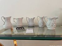 Five white porcelain miniature jugs arranged in a row on glass surface for display showing different embossed heritage designs