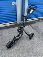 Black three-wheeled golf club caddy standing upright on asphalt with visible small compartment and brake lever on the handle.