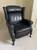 Front-facing view of the black faux leather recliner showing the wingback design, cushioned seat and back, and gold-colored studs along the armrests.