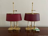 Pair of brass base desk lamps with dual white candle-style bulb holders and maroon adjustable shades