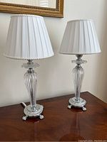 Pair of matching table lamps with glass bodies and silver plate bases, topped with white pleated shades, displayed on wooden surface.