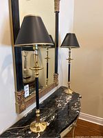 Pair of polished brass and black table lamps with black shades on a black marble-top table, reflecting in a large ornate mirror