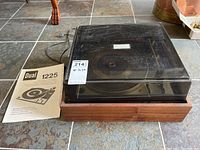Dual 1225 Hi-Fi automatic turntable with wood case and plastic cover alongside the instruction manual on a tiled floor.