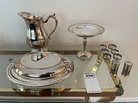 Front view of the silver plate water jug, covered serving dish, pedestal candy dish, salt and pepper shakers, and napkin rings arranged on a glass table.