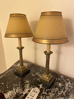 Pair of vintage French brass candlestick lamps with square dark stone bases and gold-toned fabric lampshades placed on a dark marble table against a light wall.