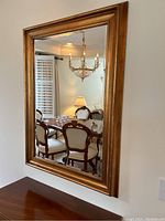 Front view of the rectangular beveled gold framed wall mirror hanging on a wall with reflection of a dining room set.