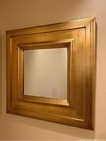 Front angled view of the square wall mirror with gold tinted wood frame showing layered molding and reflective glass surface.