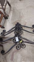 Four heavy cast iron scroll pieces with ornamental and floral designs, showing rust and dirt, laid on a concrete floor.