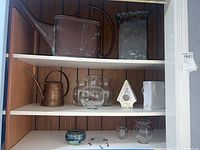 Copper watering cans including a large 2-gallon can and smaller can, assorted glass vases, ceramic vase, ceramic birdhouse, and wire dreamcatcher arranged on shelves