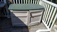 Front view of two-door plastic deck box with green lid and beige body on a porch deck.