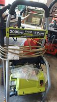 Front view showing Ryobi branding, 2000 PSI, 1.2 GPM labels, power switch, and coiled hose.