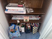 Wooden shelving unit with multiple bags containing holiday and all occasion gift wrapping supplies including bows, gift bags, wrapping paper, and gift tags.
