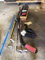 Photo showing multiple sports items on concrete floor including hockey stick, golf clubs, gloves, black bag, and ski roof rack
