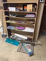 Photo showing multiple shelves filled with craft supplies including colorful corrugated papers, label makers, wall decals, folders, notepads, and a paper cutter on the floor.