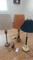 Photo of three metal-based table lamps with different colored shades on wooden floor showing overall condition and color variety.