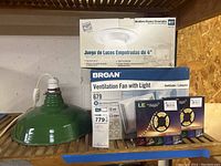 Vintage green metal hanging light with electrical cord and plug on wooden shelf next to boxed recessed light kit, Broan ventilation fan with light, and LED rope lighting strips.