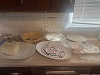 Overview of various trays, bowls, plates, glass serving dishes, and a heart-shaped ceramic dish on a countertop under kitchen cabinets.