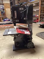 Front view of the black and gray bench top band saw showing the metal base, flat cutting table with manual and spare blades on top, and the motor attached at the bottom right.