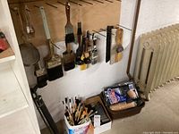 View of hung paint brushes of various sizes and types, sanding blocks below, paint rollers and brushes in plastic tub to right, radiator visible nearby.