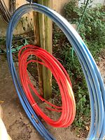 Blue and red coils of PEX tubing resting near a wooden post outdoors, with visible dirt and grass surroundings.