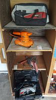 Shelf containing empty RotoZip tool bag, Orange Black and Decker sander, Milwaukee drill, and black plastic circular saw case.